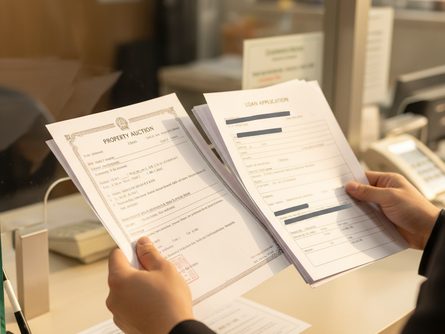 손으로 잡은 부동산 경매 낙찰결정문과 대출 신청 서류 클로즈업, 흐릿한 은행 창구 배경